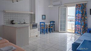 A compact hotel room with a kitchenette, a dining area with blue chairs, a sofa, and a view through a sliding door to the outside. - Enplo Apartments (Hersonissos)