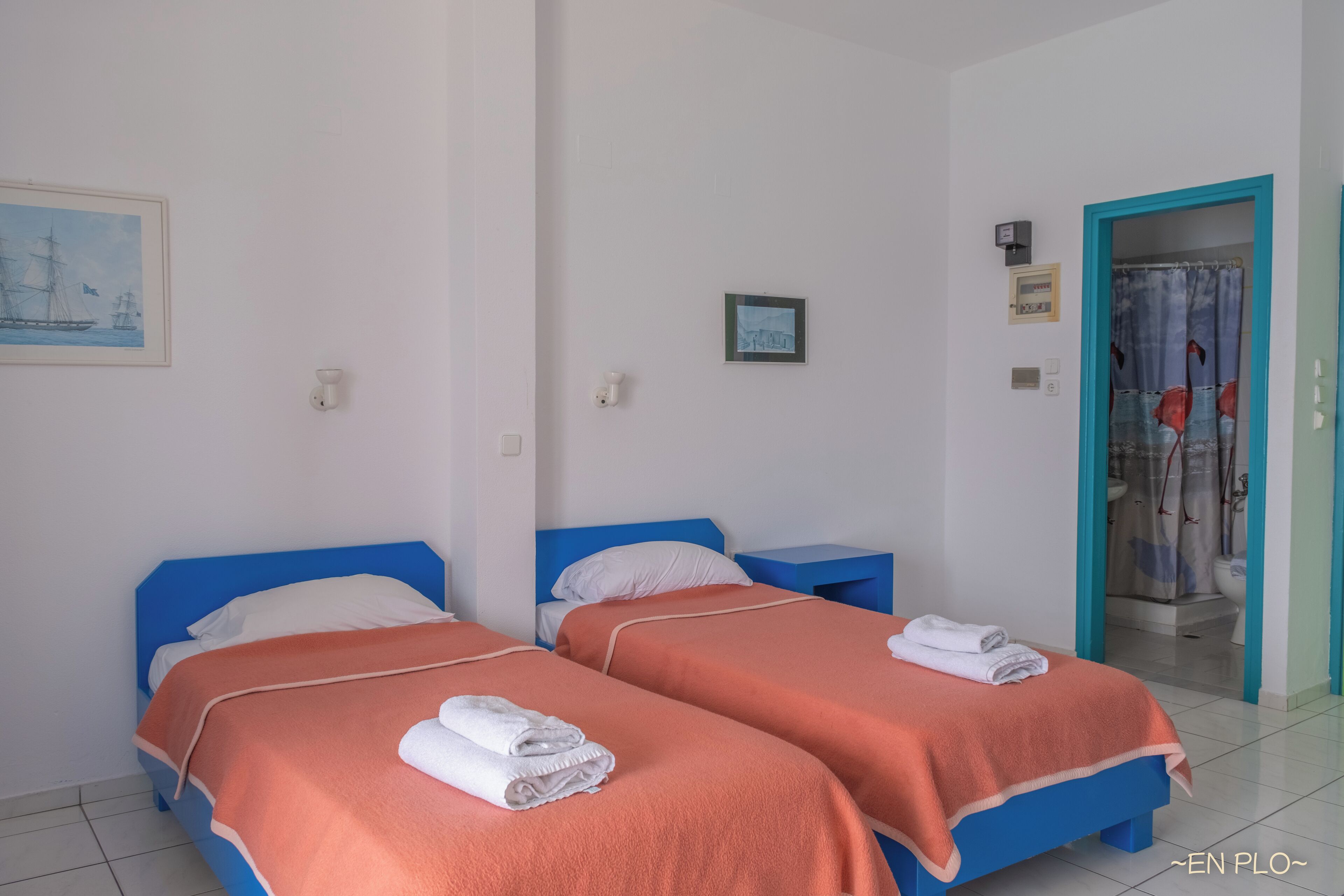 Een hotelkamer met twee bedden, elk met een blauwe hoofdbord en oranje beddengoed. Er liggen witte handdoeken op de bedden en een badkamer met een douchegordijn.