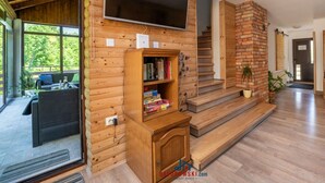 L'intérieur d'un chalet moderne en bois, doté d'un téléviseur à écran plat fixé au-dessus d'une bibliothèque en bois, d'un escalier menant à un étage supérieur et offrant une vue sur un jardin luxuriant grâce à de grandes fenêtres.
