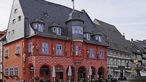 A red building with multiple windows and a corner turret, featuring outdoor seating and a fountain. - Granetal.quartier-apartment Harzidylle (Goslar)