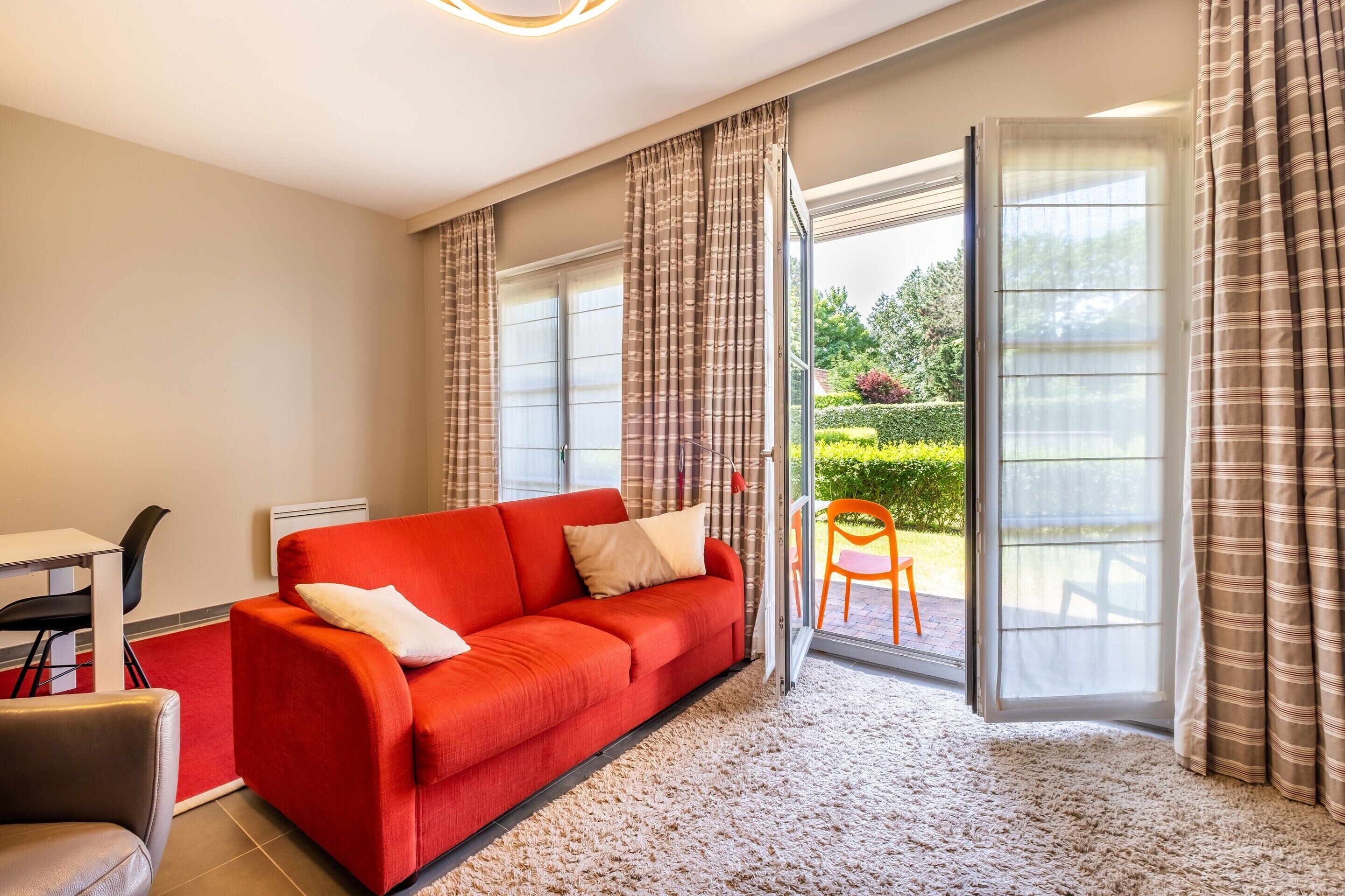 A modern living room with a red sofa, a dining table with chairs, and a view of a garden through a sliding glass door.