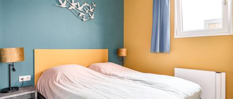 A bedroom with a bed, a nightstand, a lamp, a wall art piece, a window with curtains, and a radiator.