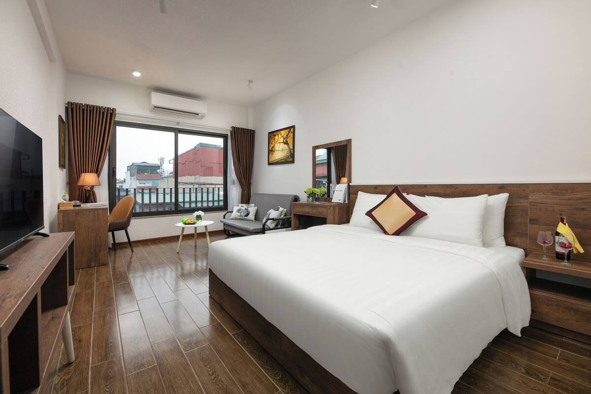 A hotel room with a large bed, a desk with a chair, a TV, a small table with a plant, and a window with curtains.
