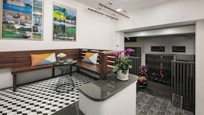 Une salle d’attente avec un sol à carreaux, un banc en bois avec des coussins, une petite table sur laquelle est posé un vase de fleurs, et deux affiches accrochées au mur.