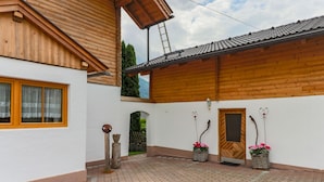 A wooden building with a tiled roof, a window with a wooden frame, and a door with a wooden panel. There are two potted plants with flowers. - Cosy Holiday Home Near ski Area (Piesendorf)