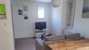 A spacious living area with a wooden dining table, a grey sofa, and a television in the background.