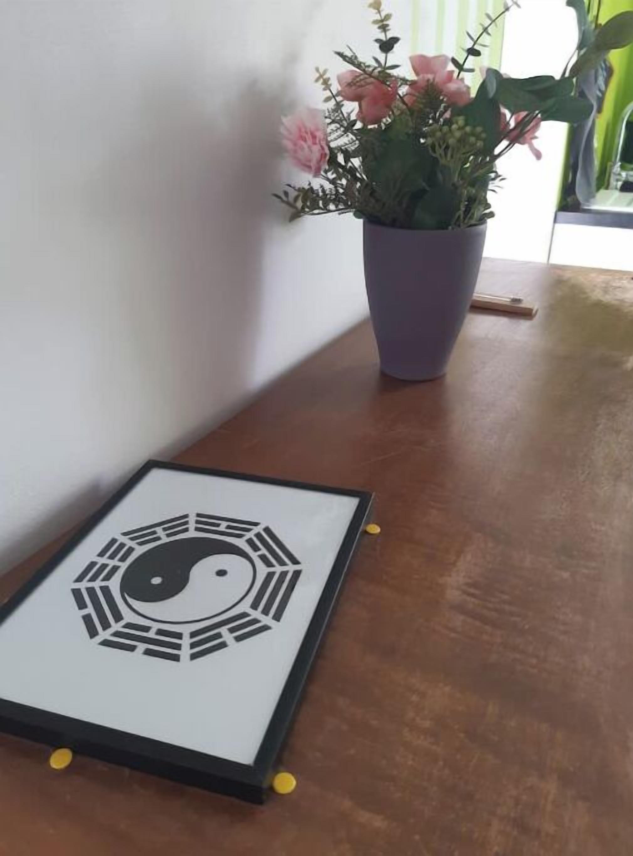 Un símbolo ying yang enmarcado sobre una mesa de madera, junto a una maceta con flores rosas.