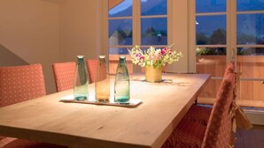 A dining table with three glass bottles and a flower vase, set against a backdrop of large windows with a mountain view. - Plaatsje in het Mesnerhuis (Lenggries)