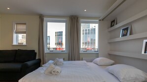 A hotel room with a large bed, a dark-colored sofa, and two windows offering a view of buildings. - Apartment in Vlissingen Near the Beach (Vlissingen)