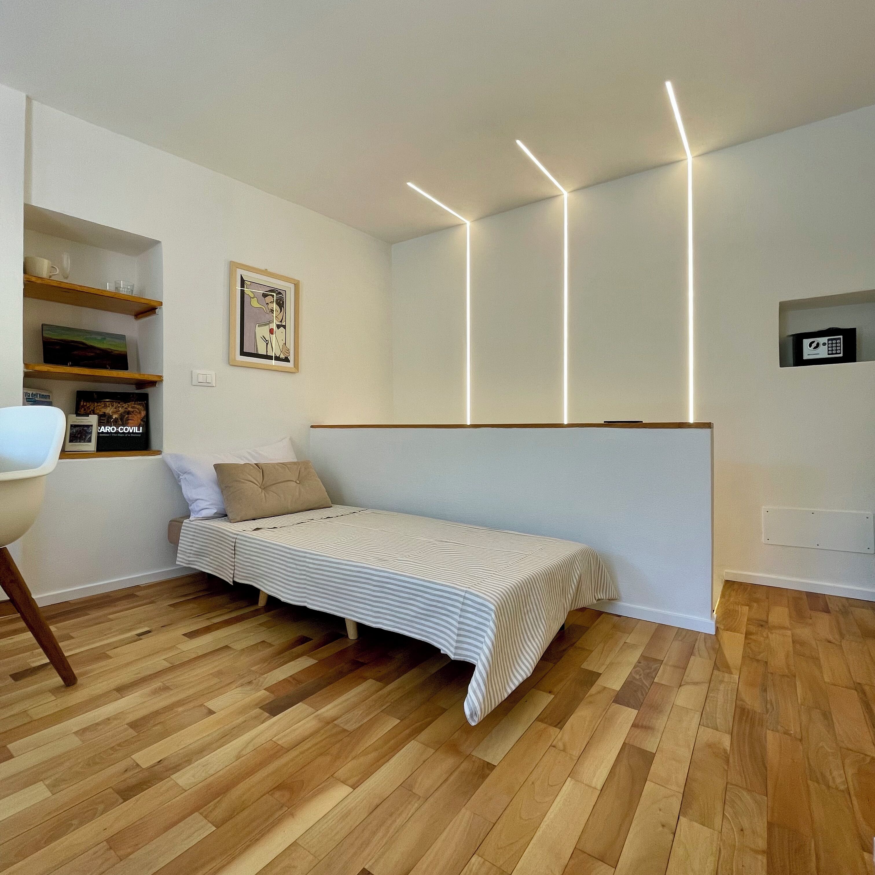 A minimalist bedroom with a bed, wooden flooring, and modern lighting.