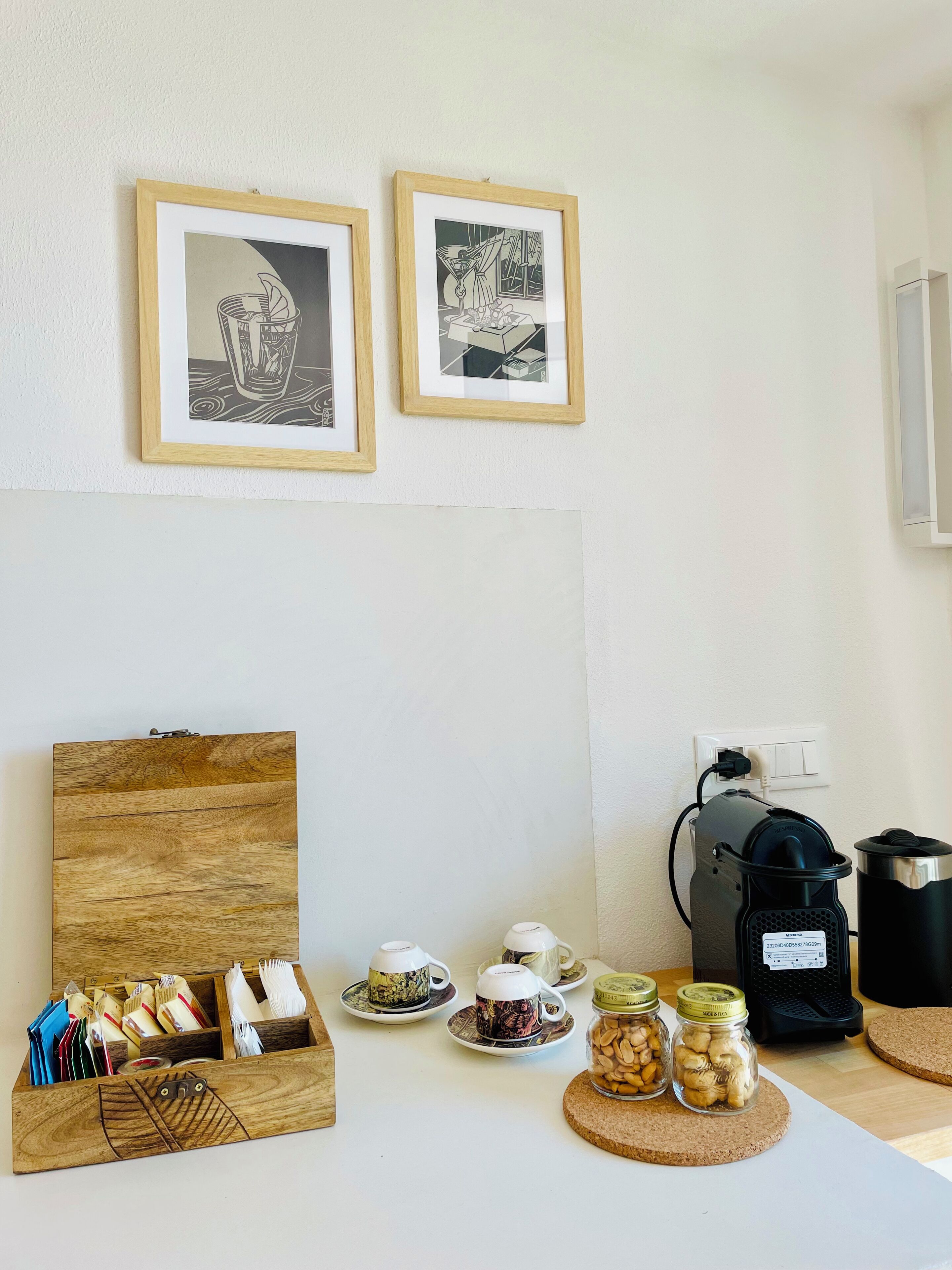 A room with two framed black and white artworks on the wall, a wooden sideboard with tea sets and jars, and a coffee machine on the table.