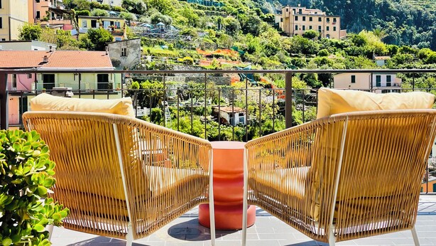 Een terras met rieten stoelen en een rode tafel, met uitzicht op een heuvel met gebouwen en groen.