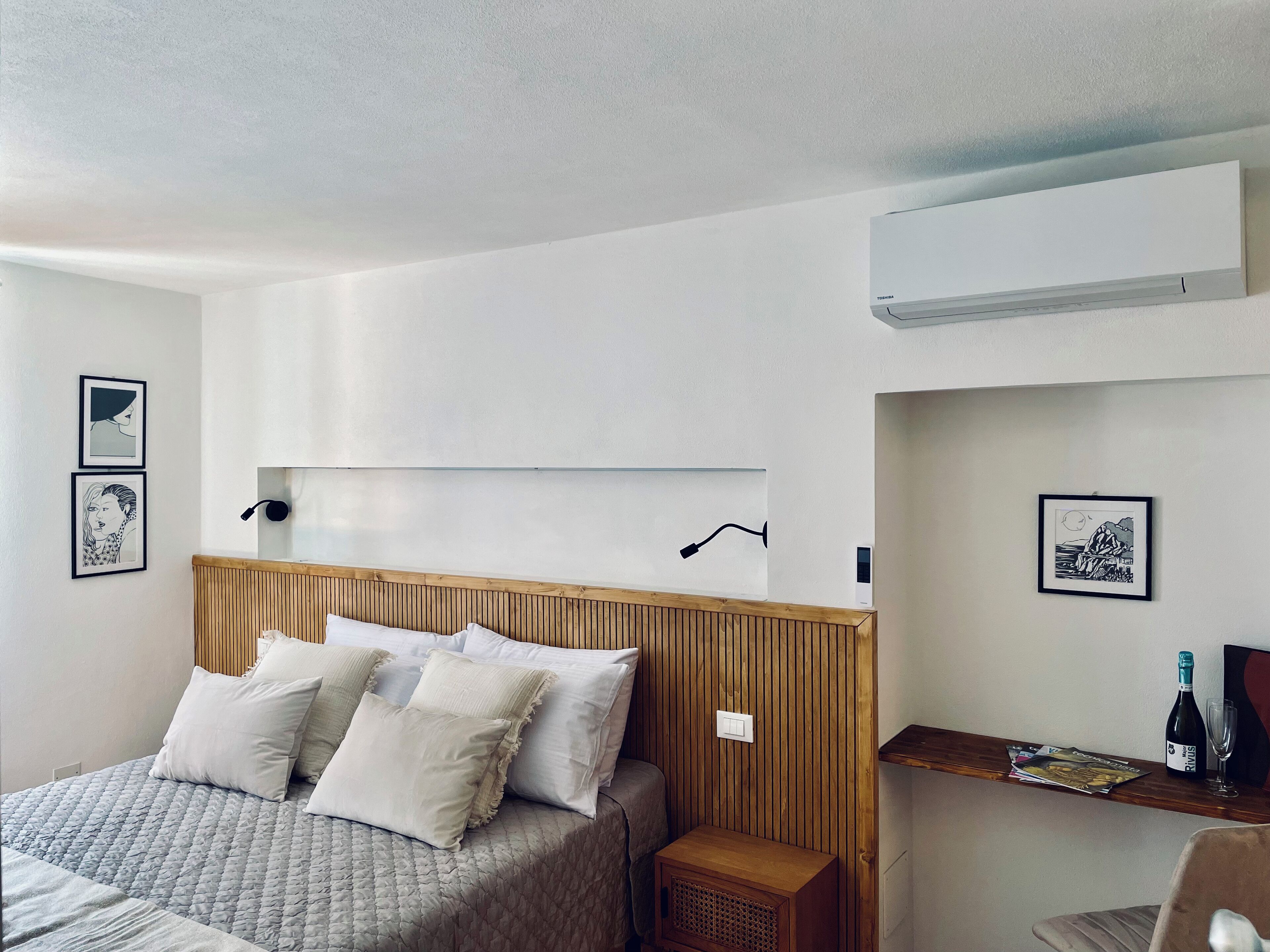 Un dormitorio moderno con cabecera de madera, cama con ropa de cama blanca y gris, mesita de noche con lámpara y dos cuadros enmarcados en la pared.