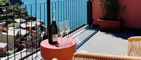 Un balcón con sillas de mimbre, una mesa con una botella y vasos, y vista al mar.