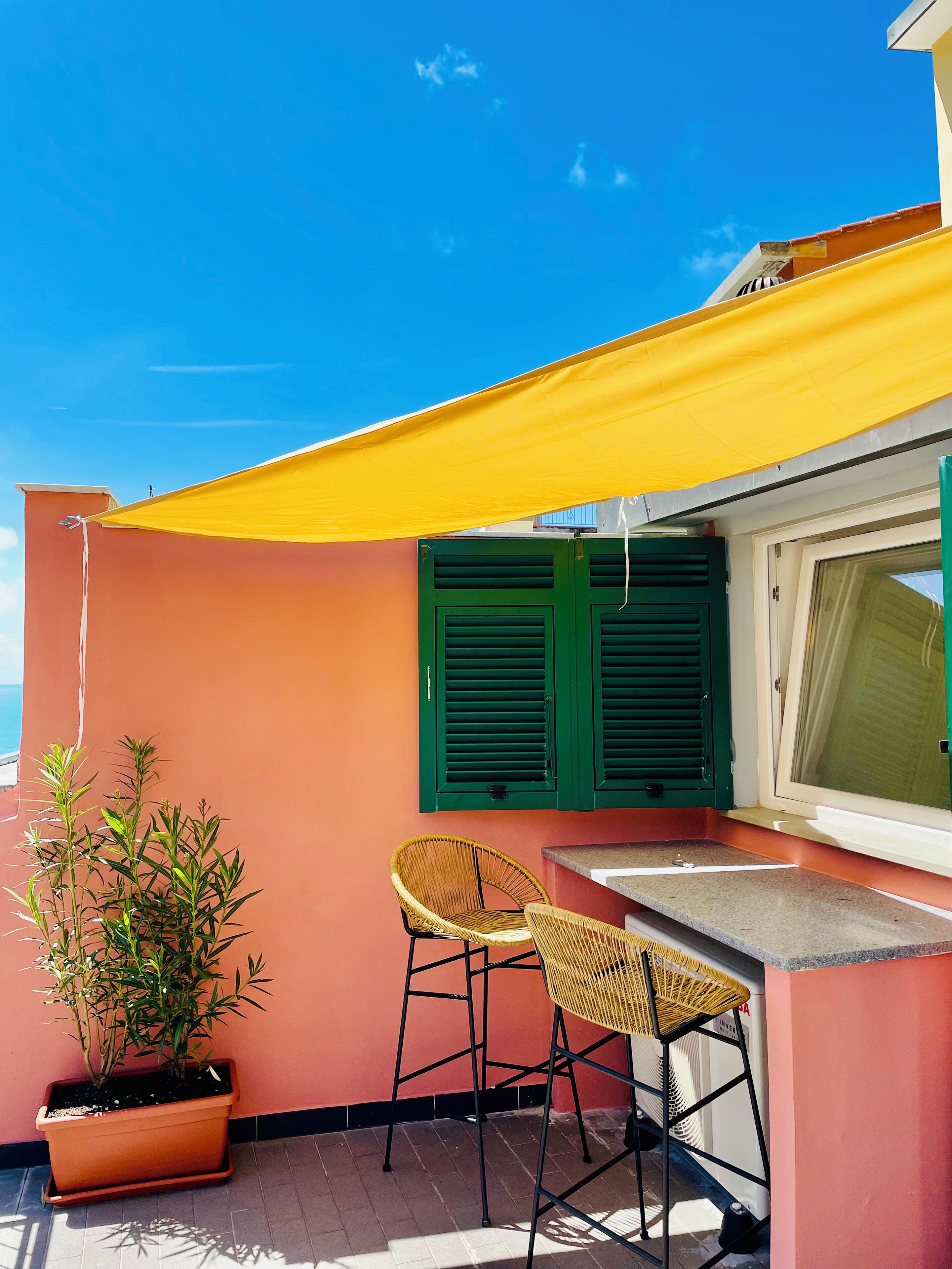A small outdoor seating area with a table and two chairs, a potted plant, and a yellow awning providing shade.