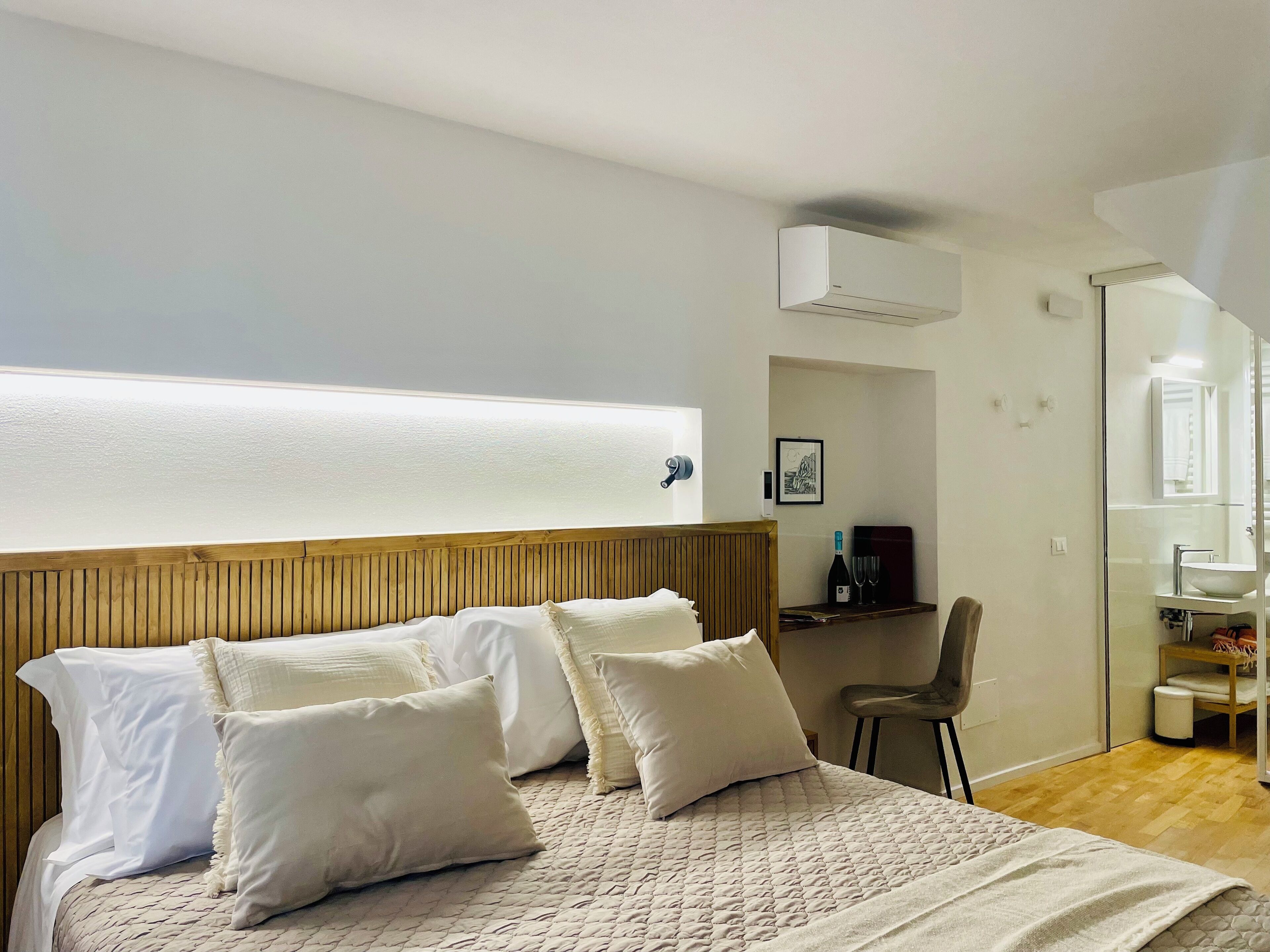 A bedroom with a wooden headboard, a bed with white bedding, a desk with a chair, and a bathroom with a mirror and sink.