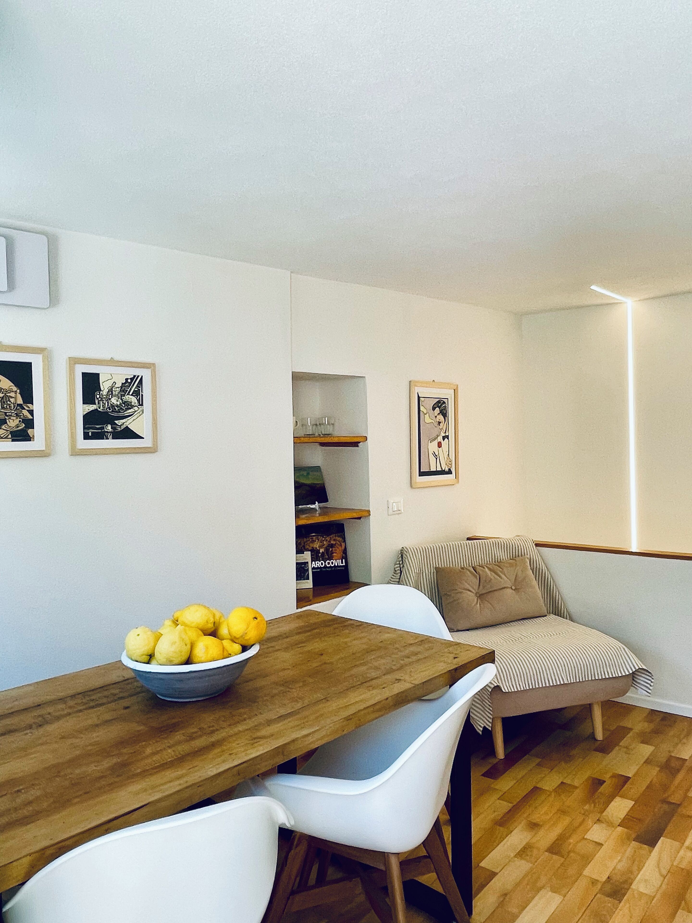 Un comedor con una mesa de madera, sillas blancas y un cuenco de limones. Hay cuadros en la pared y una silla a rayas.