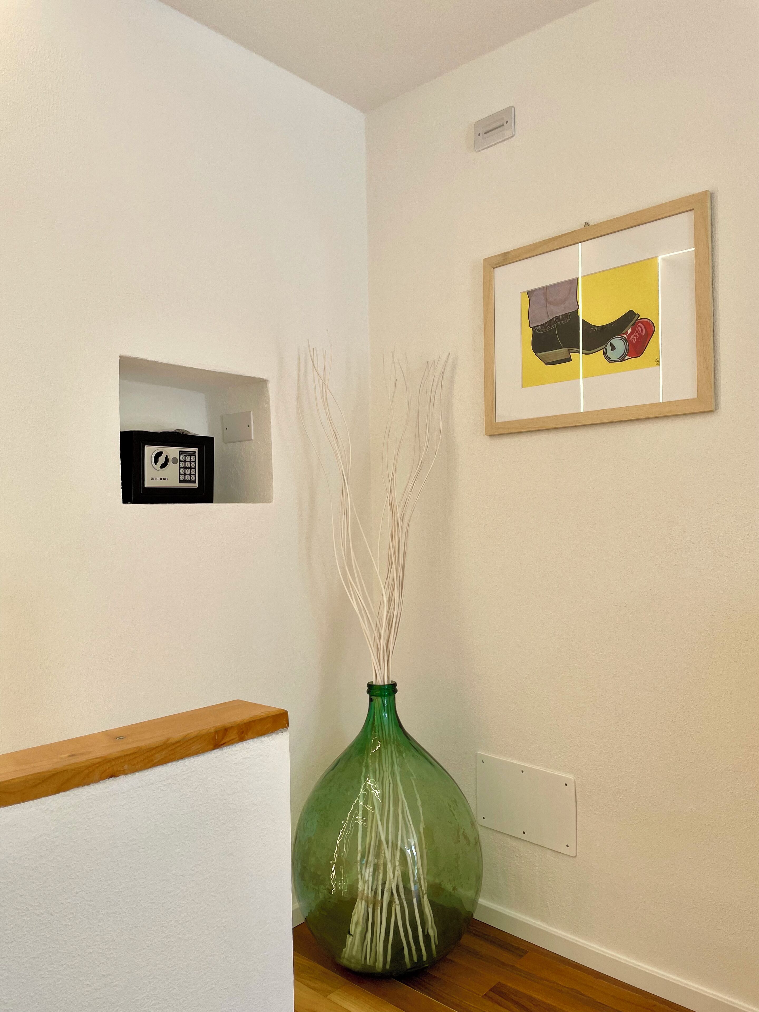 A room with a green vase with branches, a framed picture on the wall, and a small black device on the wall.