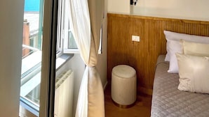 Een hotelkamer met een bed, een klein krukje, een houten hoofdeinde en een raam met gordijnen.