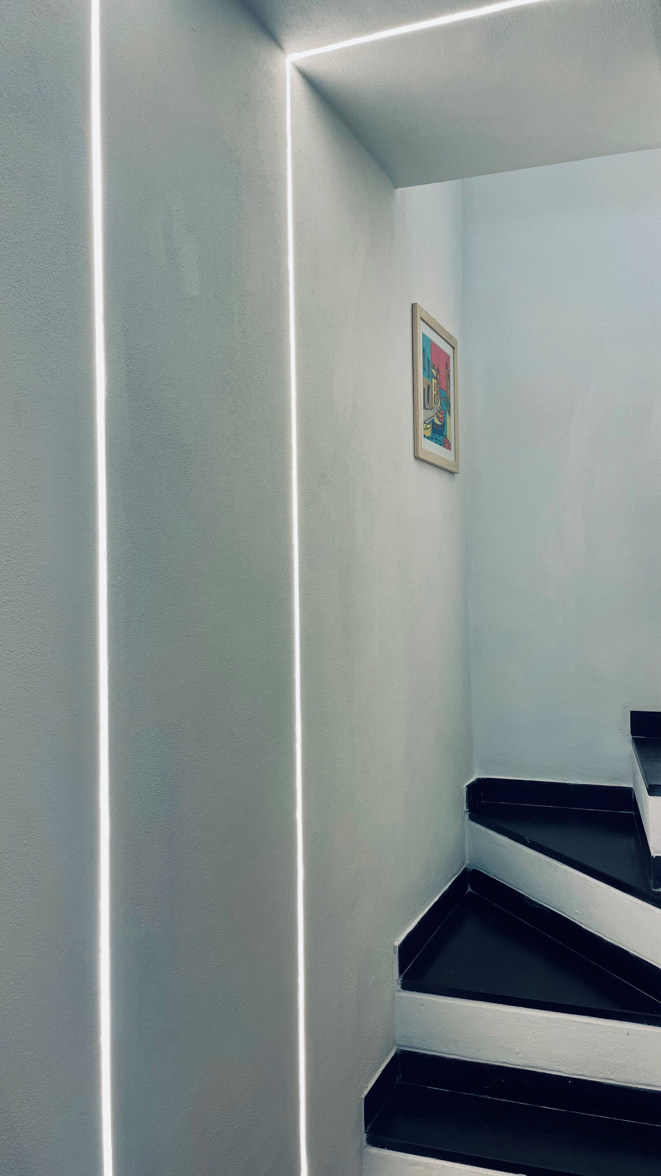 A corner of a room with a white wall, a framed picture, and a black staircase.