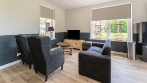 Un salon moderne comprenant un canapé, un fauteuil et un téléviseur, de grandes fenêtres et un coin cuisine avec un réfrigérateur.