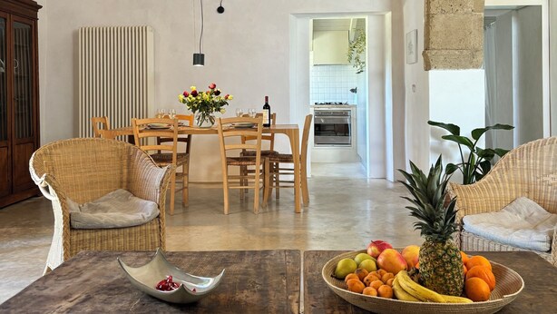 Un vaste espace de vie doté d’un haut plafond en bois, d’une table à manger avec des chaises, d’un canapé en osier et d’un panier de fruits posé sur une table en bois.