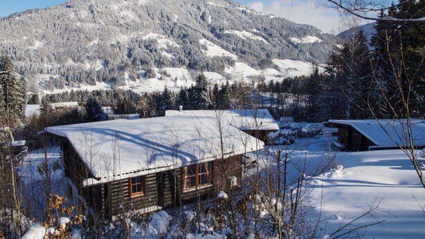 A wooden cabin with a snow-covered roof, surrounded by a snowy landscape and mountains in the background. - Wastl Comfortable Holiday Residence (Woergl)