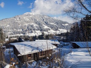 Une cabane en bois, dont le toit est recouvert de neige, entourée d’un paysage enneigé et avec des montagnes en arrière-plan.
