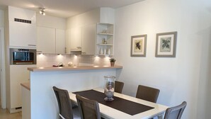 A modern kitchen with white cabinets, a built-in oven, and a dining area with a white table and wicker chairs. - Apartment in Nieuwpoort by the Beach (Nieuwpoort)