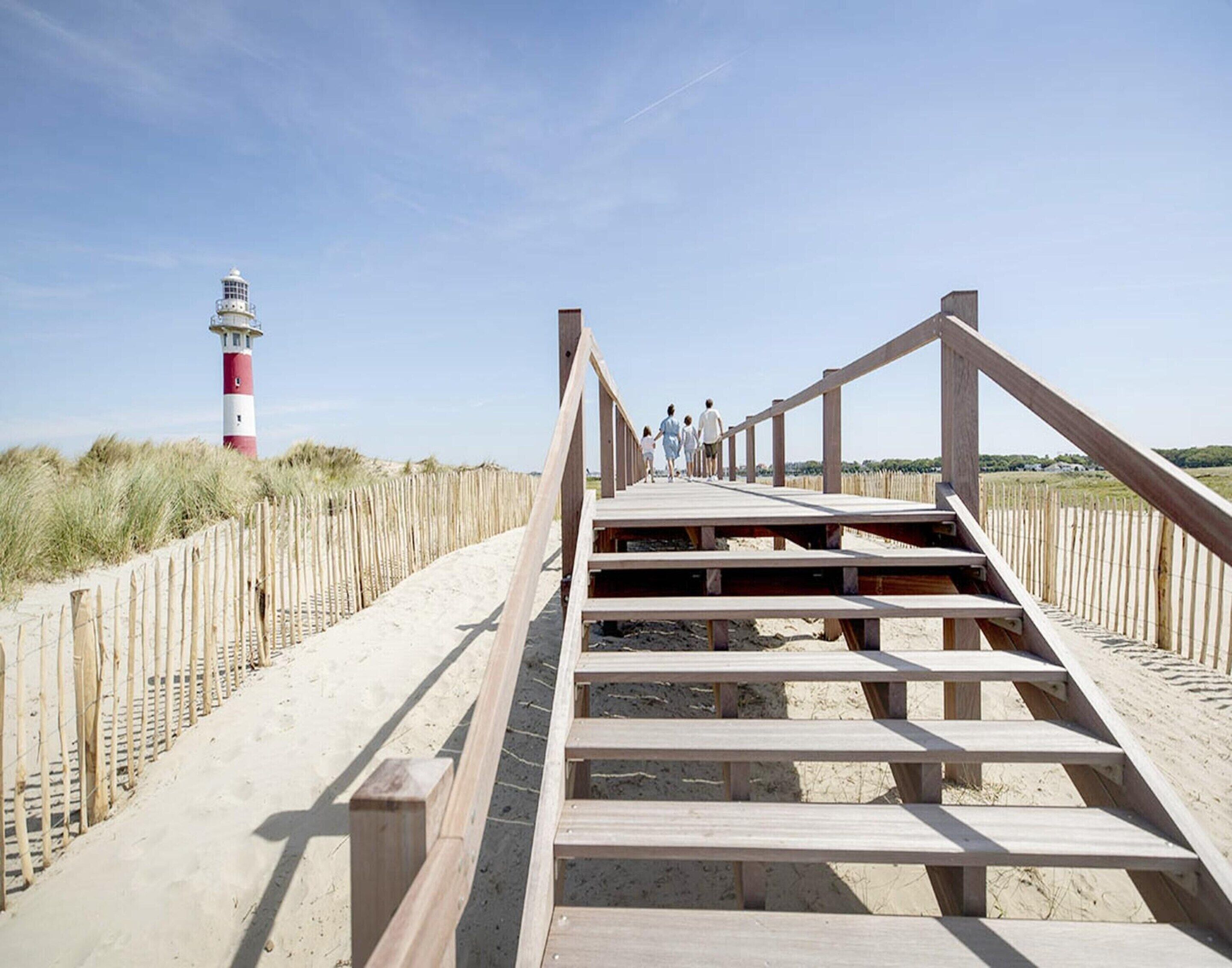 A wooden boardwalk leading to a lighthouse, with a sandy beach and grassy dunes on either side.