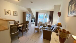 A modern living room with a dining area, a sofa set, a TV, and a large window with curtains. - Apartment in Nieuwpoort by the Beach (Nieuwpoort)