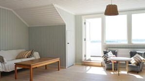 Quarto com piso de madeira, um sofá branco, uma mesa de madeira e um sofá cinza com almofadas listradas. Há uma grande janela com vista para o exterior.