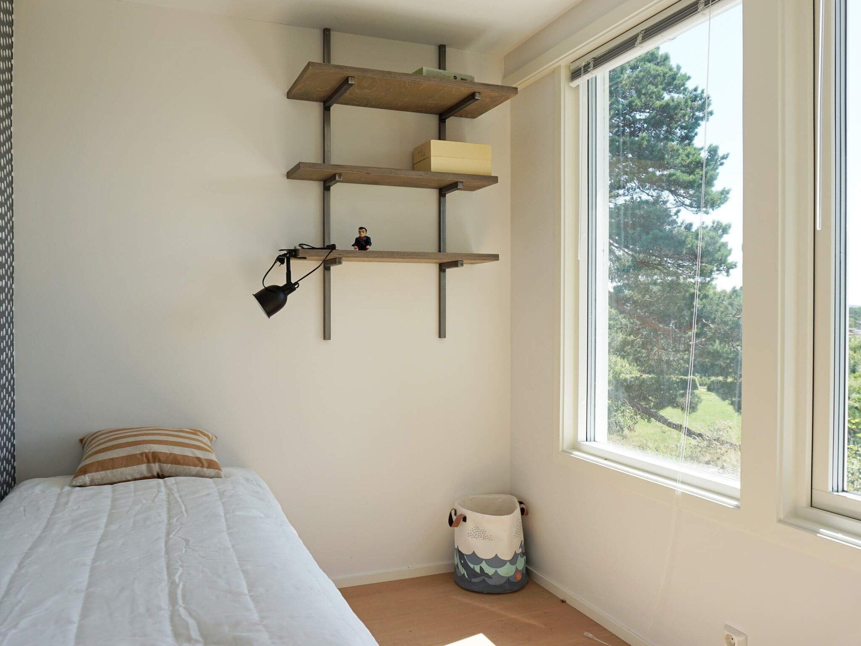 Un dormitorio con cama, ventana con vista a árboles y estante con lámpara.
