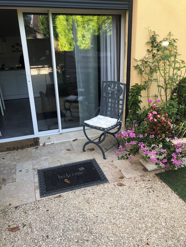 Un patio con una silla, un felpudo y un pequeño jardín con flores rosas.