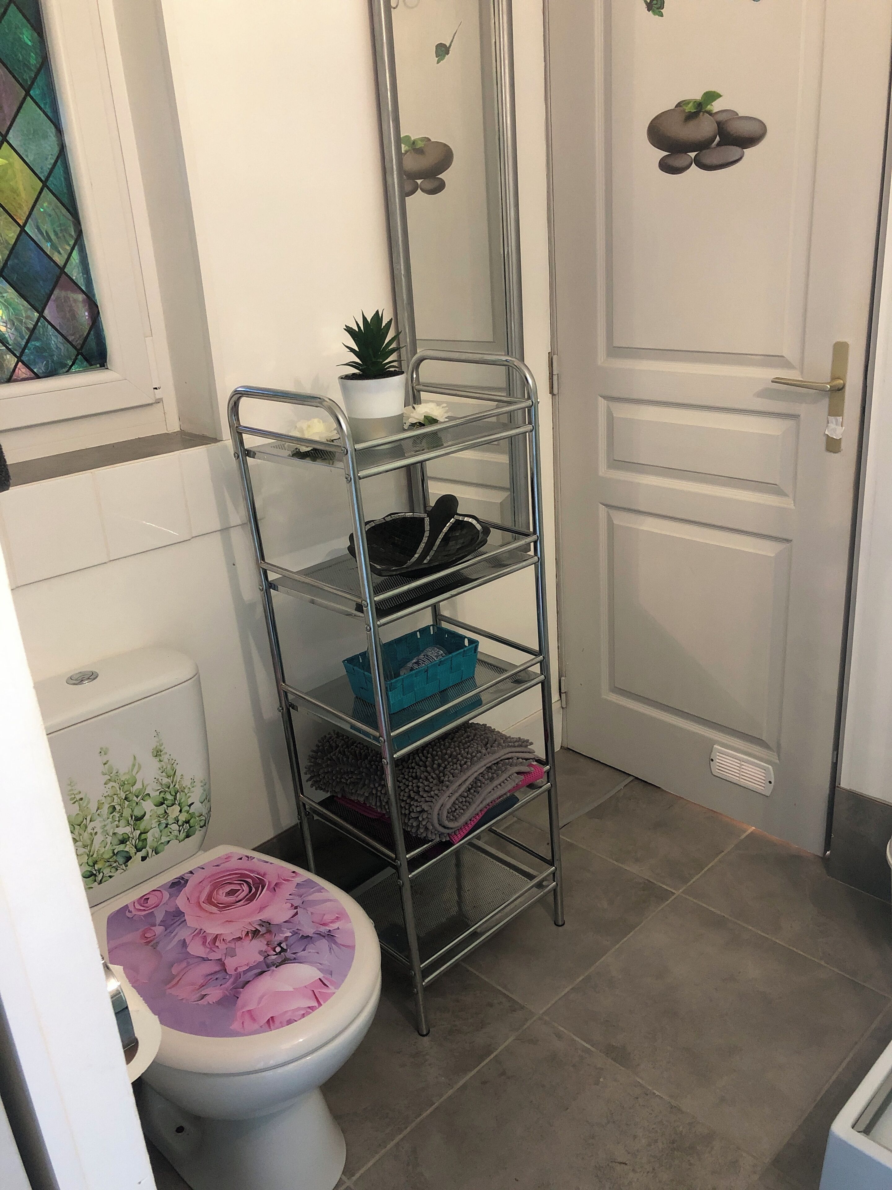 Un baño con un inodoro, un estante metálico con diversos objetos, una planta en maceta y una puerta con diseño de piedra.
