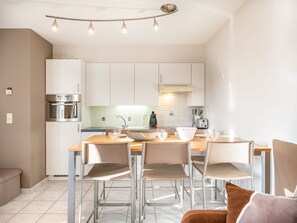 A modern kitchen with white cabinets, a built-in oven, and a dining area with four chairs and a wooden table. - Apartment in Koksijde Near the Sea (Koksijde)