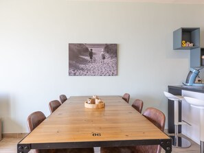 Een vergaderruimte met een houten tafel, acht stoelen, een ingelijst fotoportret aan de muur en een kitchenette met een koffieapparaat en opslagplanken.