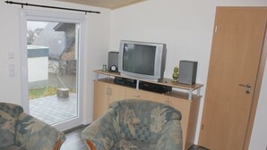 Sala de estar com televisão sobre suporte de madeira, duas poltronas estampadas e uma porta de vidro deslizante para área externa.
