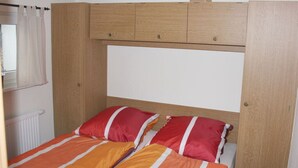 Uma cama de casal com duas almofadas, cabeceira de madeira e janela com cortinas.