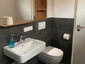 Een badkamer met een wit wastafel, toilet en spiegel boven het wastafel. Er is een plankje met een rol toiletpapier en een fles handzeep.