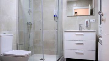 A modern bathroom with a glass-enclosed shower, a white toilet, and a vanity with a mirror and a soap dispenser.