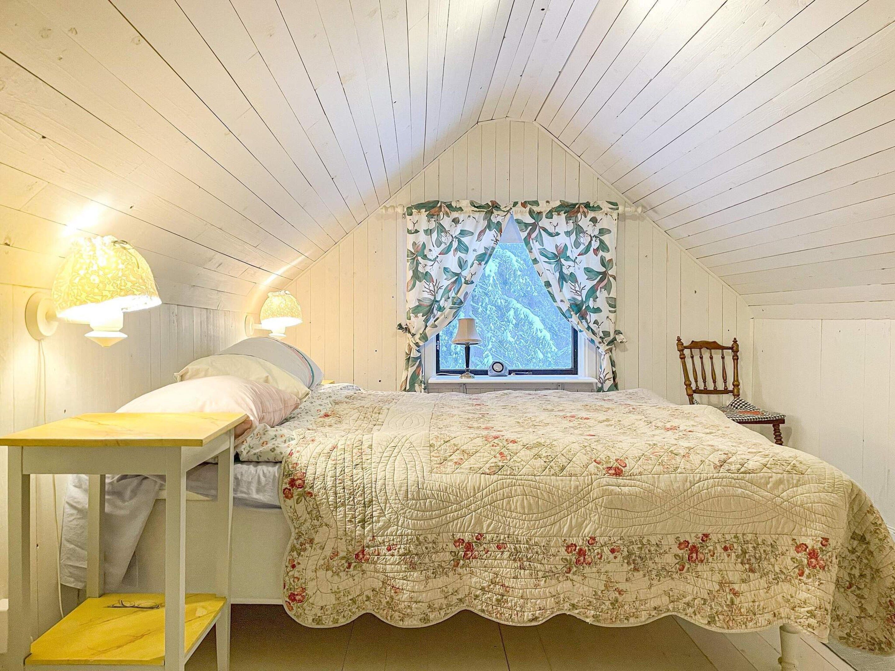 Un dormitorio con techo inclinado, una cama con colcha floral, dos lámparas de pared, una ventana con cortina floral y una silla de madera.