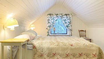 Un dormitorio con techo inclinado, una cama con colcha floral, dos lámparas de pared, una ventana con cortina floral y una silla de madera.