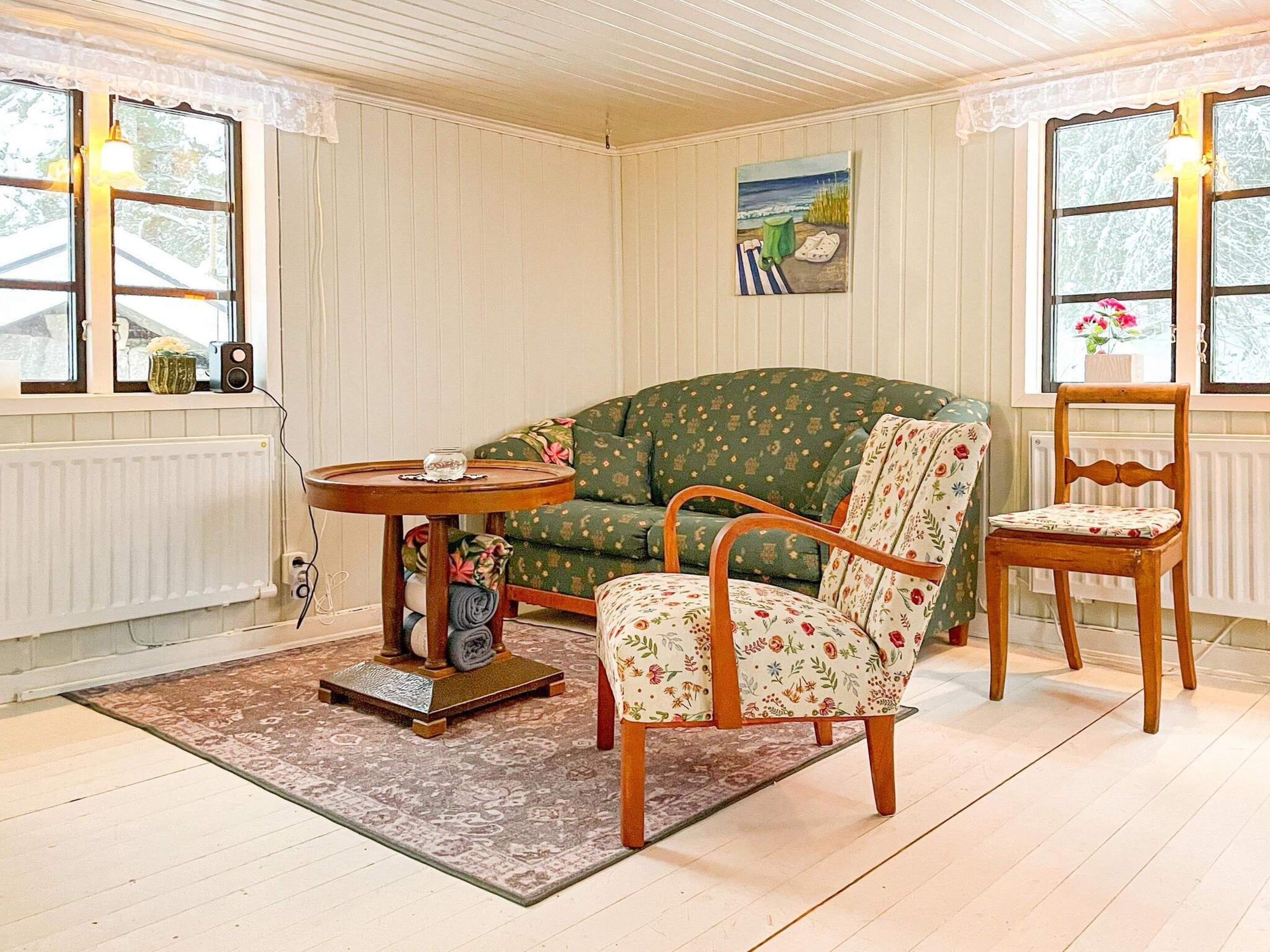 A room with a green patterned sofa, a wooden chair, a small table with a glass object, and a painting on the wall.