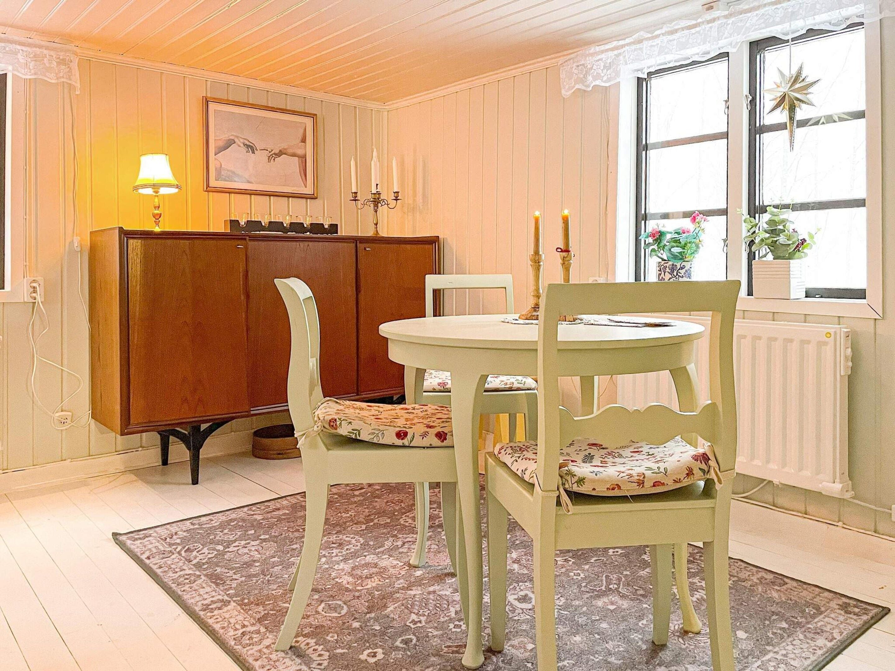 A dining area with a wooden cabinet, two chairs with floral cushions, a table, and a window with plants.