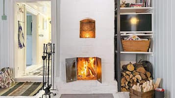 Una habitación con chimenea de ladrillo blanco, un televisor sobre una estantería y una puerta que da a otra habitación.