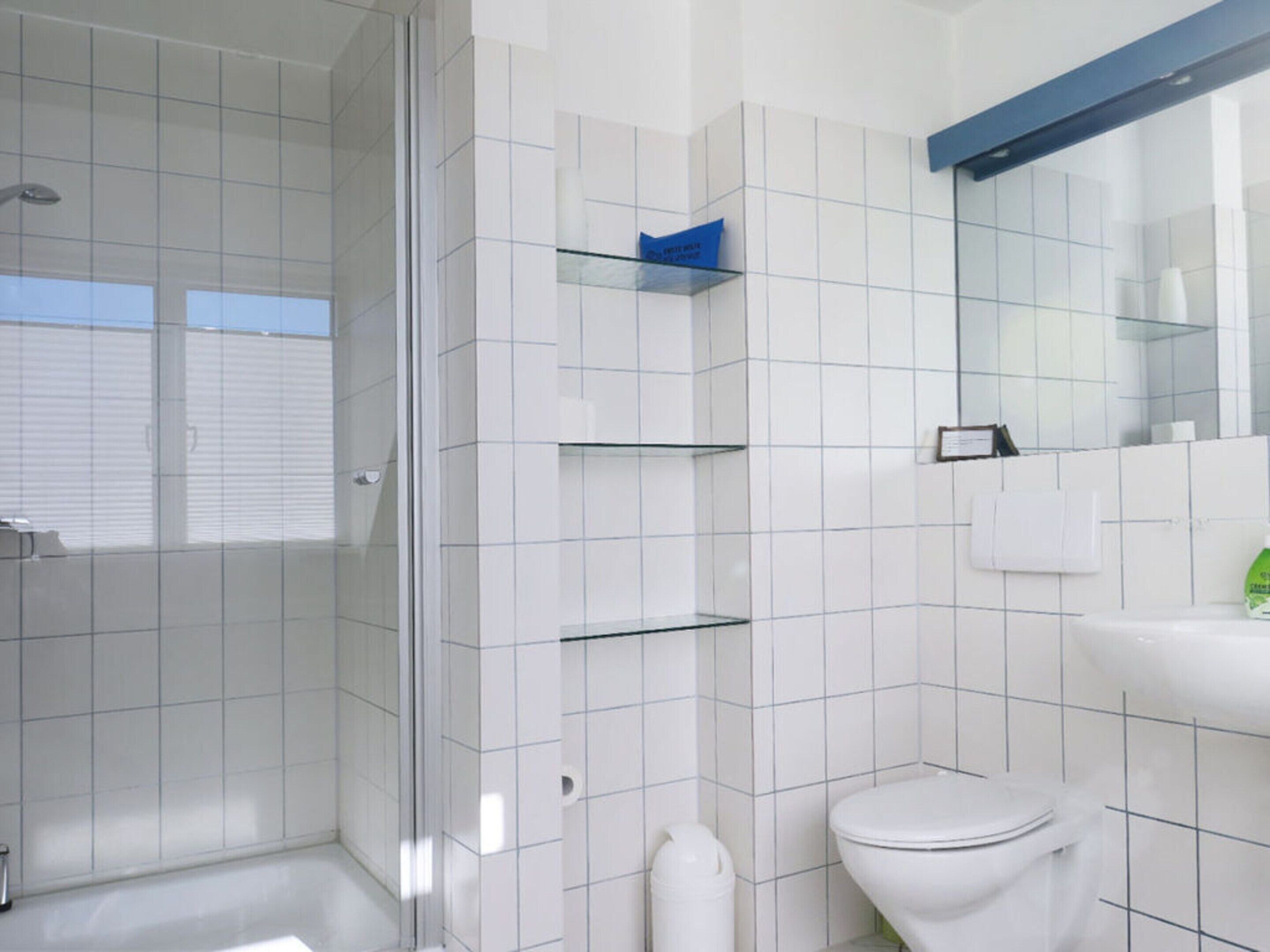 Un baño moderno con azulejos blancos, cabina de ducha de vidrio, inodoro y lavamanos con espejo superior.