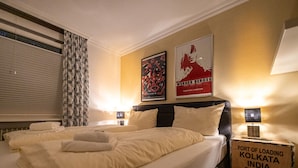 A hotel room with a large bed, two bedside tables, a window with blinds, patterned curtains, and two framed posters on the wall. - Hexenzauber Hahnenklee (Goslar)