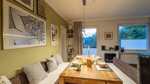 A cozy living room with a wooden dining table, a sofa, and a bookshelf. There are framed artworks on the wall and a window with a view of greenery. - Hexenzauber Hahnenklee (Goslar)