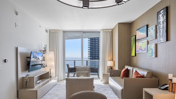 A modern hotel room with a balcony, a flat-screen TV, a sofa, a dining table, and artwork on the walls.